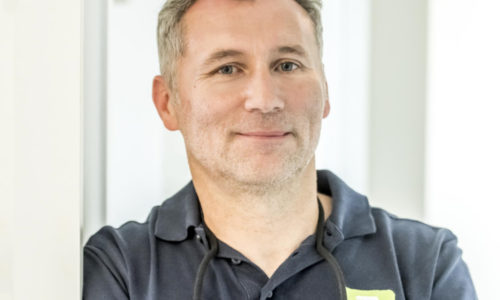 Dr Torsten Keller hat graumelierte kurze Haare und lächelt leicht. er trägt ein dunkelblaues Polohemd und lehnt sich an die weiße Wand.