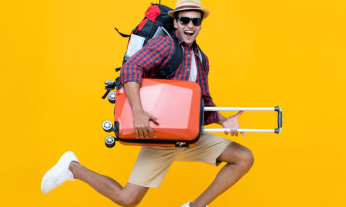 zahnpflege im urlaub - mann mit rollkoffer, hut, sonnenbrille und rucksack springt hoch und lacht. Er trägt eine weißes Tshirt, ein rotblau kariertes Hemd, eine beige Bermuda-Shorts und weiße Sneaker.