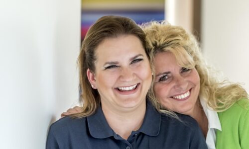 Links im Bild ist eine brünette Frau mit einem dunkelblauen Polohemd zu sehen. Sie lächelt in die Kamera. Rechts im Bild lehnt sich eine lachende blonde Frau auf Ihre Schulter. Sie trägt ein weißes Hemd und eine hellgrüne Jacke.
