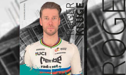 ein schlanker mann mit dunkelblonden kurzen haren, blauen augen und einem weißen radrenntrikot mit verschiedenen schriftzügen und logos schaut den betrachter an. Es ist der Radrennprofi Roger Kluge.