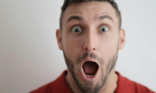 junger mann mit kurzen haaren und dreitagebart ist überrascht und öffnet augen und mund weit