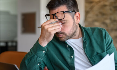 Müder Mann sitzt am Schreibtisch und fasst sich an die Nasenwurzel und seine Brille rutscht nach oben.