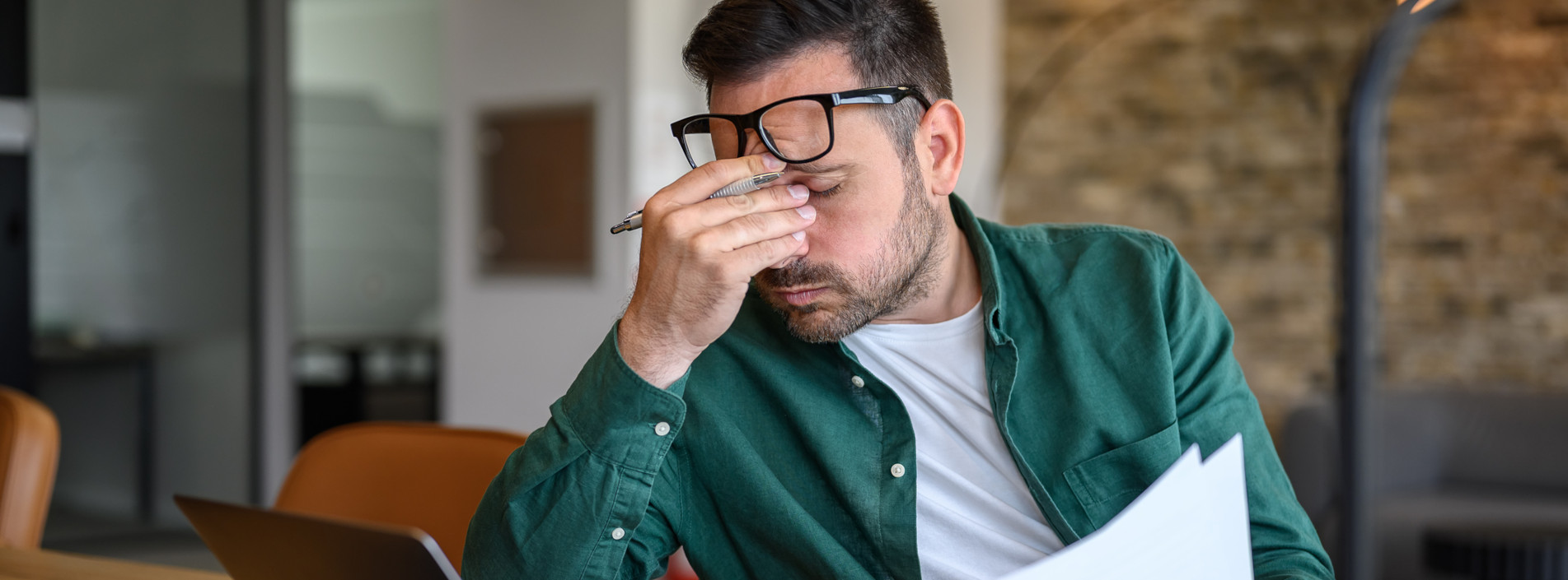 Müder Mann sitzt am Schreibtisch und fasst sich an die Nasenwurzel und seine Brille rutscht nach oben.